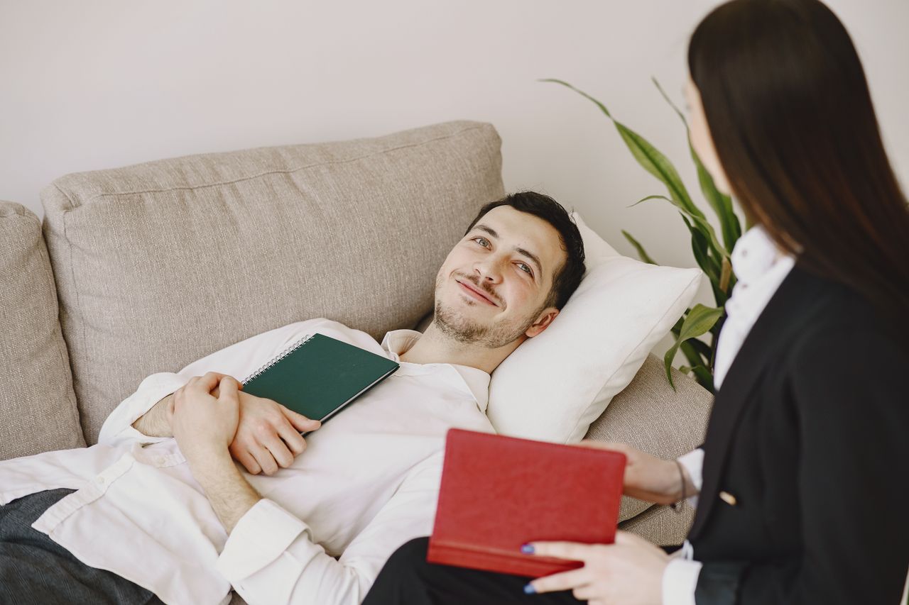 Positive man in formal wear lying on sofa with notebook in hands while smiling and talking with therapist in black suit